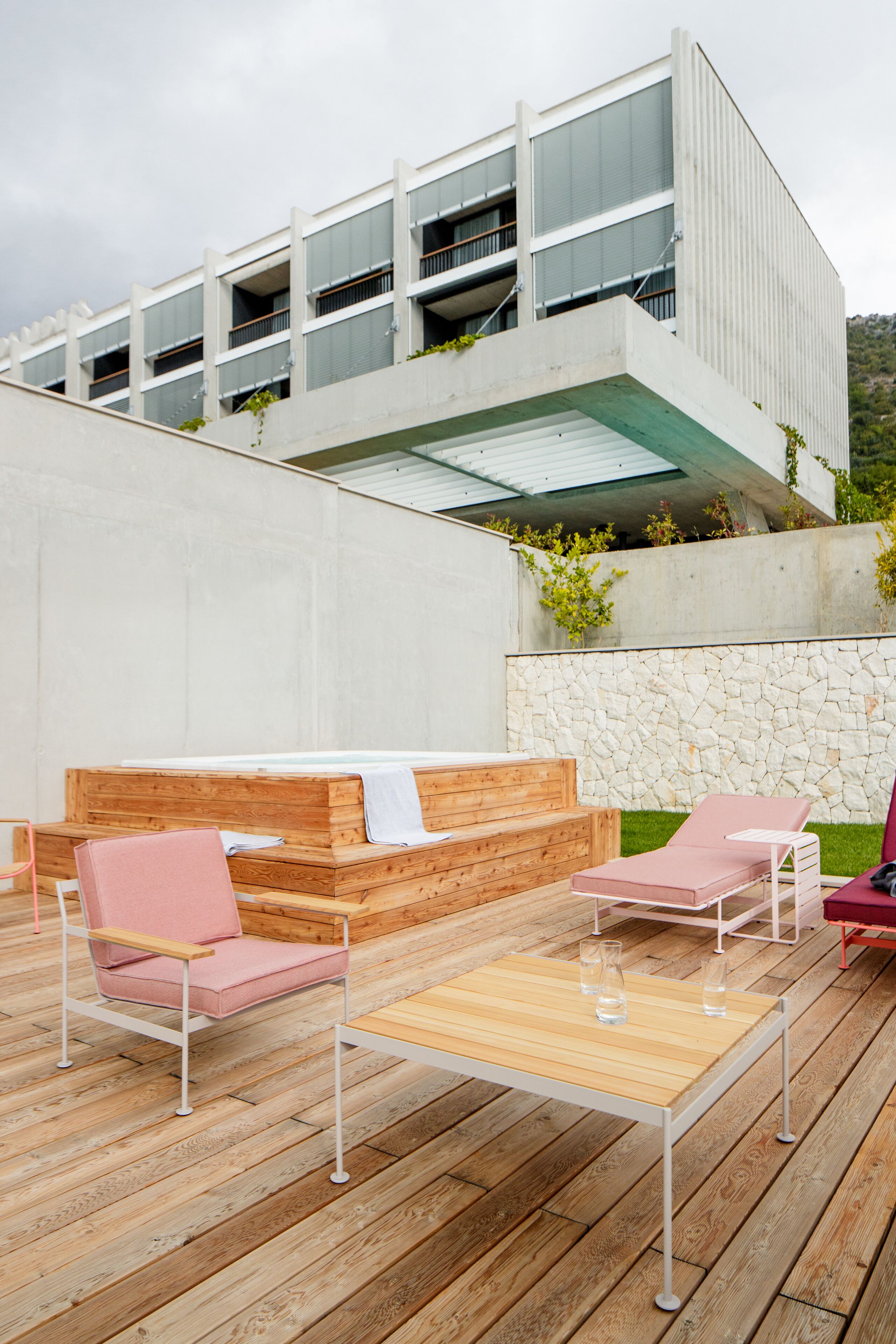 Un área de estar al aire libre con una mesa de madera y dos sillas, un muro de piedra y un edificio moderno con balcones al fondo.