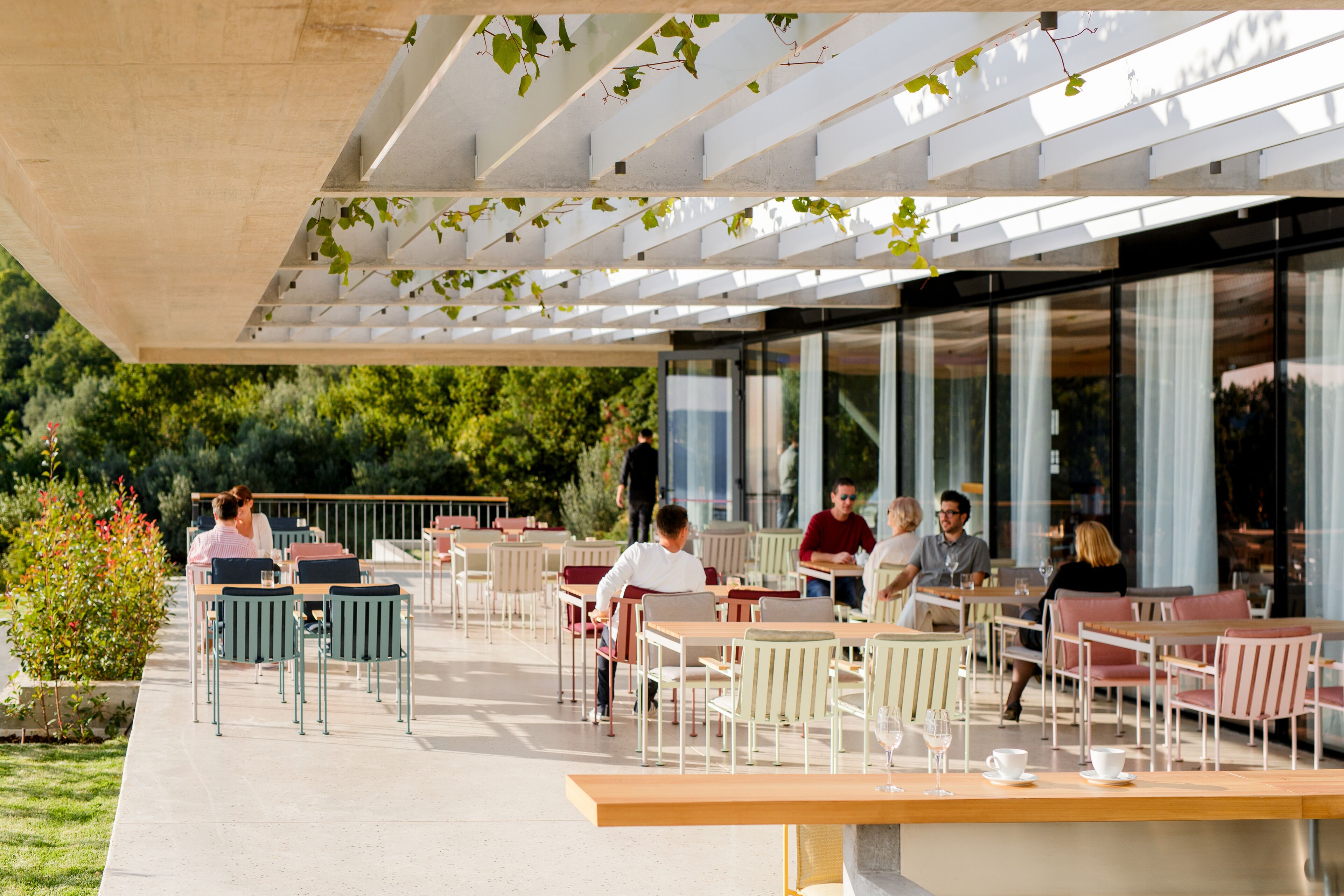 An outdoor café with tables and chairs, surrounded by greenery and large windows.
