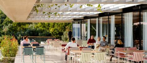 An outdoor café with tables and chairs, surrounded by greenery and large windows.