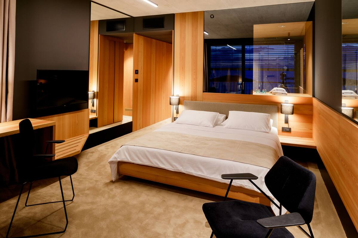 A modern hotel room with a large bed, a desk, a chair, a TV, and a window with a view.