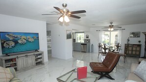 A spacious living room with a flat-screen TV, a glass coffee table, a leather armchair, and a sofa. There is a ceiling fan, a dining area with a table and chairs, and a kitchen in the background.