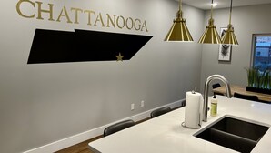 Une cuisine moderne avec un plan de travail blanc, un évier en acier inoxydable et des luminaires suspendus. Le mur est orné d’une enseigne dorée sur laquelle on peut lire « CHATTANOOGA », avec une étoile en dessous.
