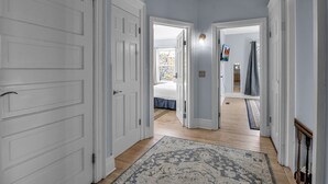 A hallway with white doors, a patterned rug, and a view into a bedroom with a bed and a window. - Spacious Historical Home by Parks, Shops, 4bd 2ba Save Weekly & monthly. (Buffalo)
