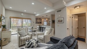 A spacious living room with a large window, a dining area, and a kitchen in the background.