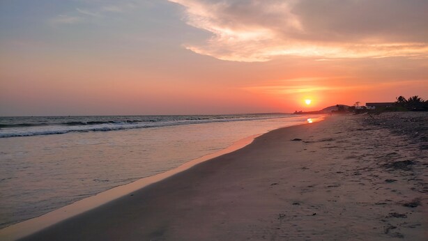 Une plage au coucher du soleil, avec une mer calme et des bâtiments qui se dessinent à l’horizon.