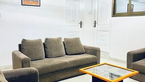 Un salon avec un canapé beige, deux fauteuils et une table basse avec un plateau en verre. Il y a deux portes blanches et un tableau encadré au mur.
