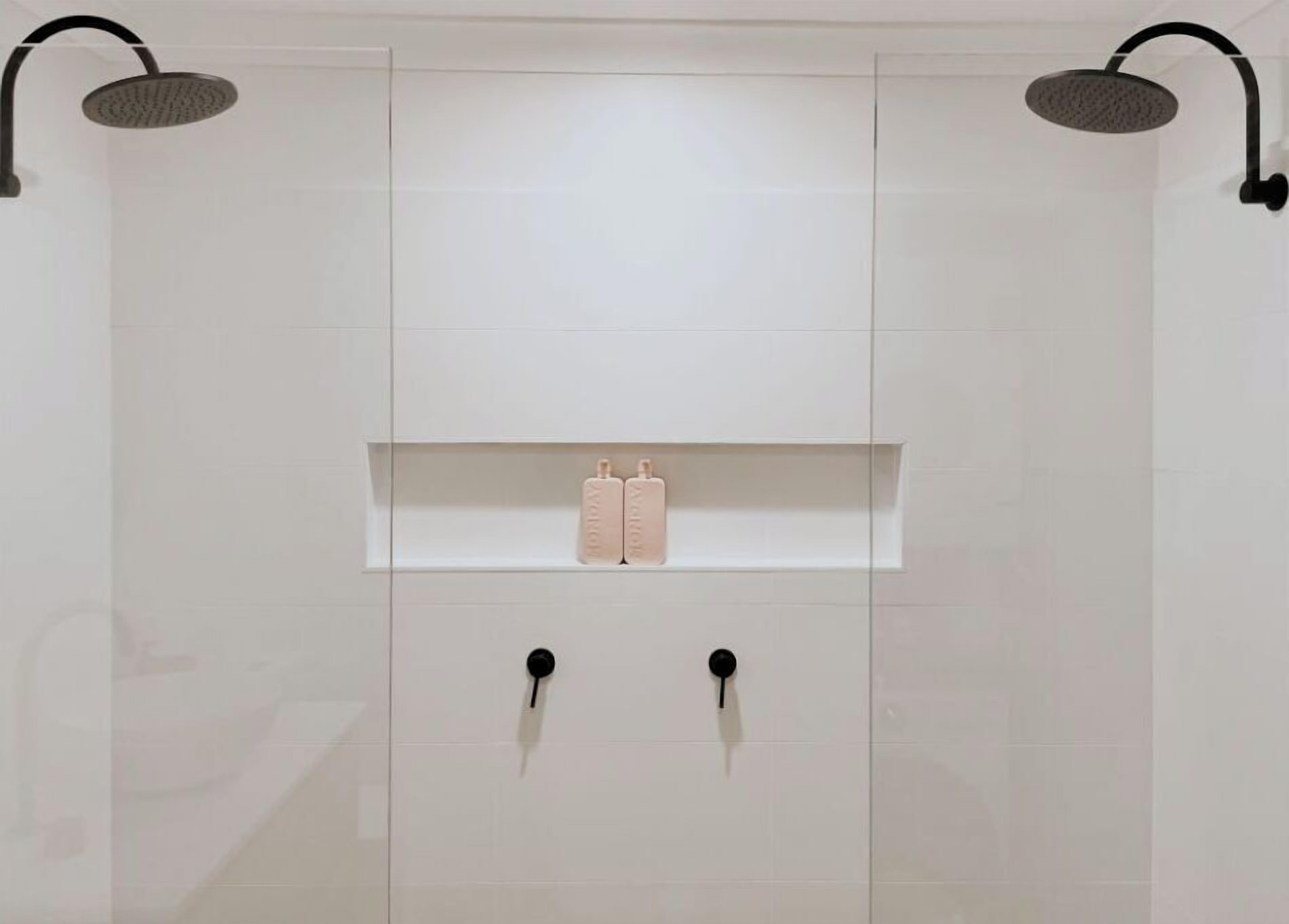 A modern bathroom with a white tiled wall, two black showerheads, and a shelf with two bottles.