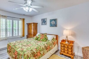 Quarto com uma cama, mesa de cabeceira de madeira, ventilador de teto e duas obras de arte penduradas na parede.