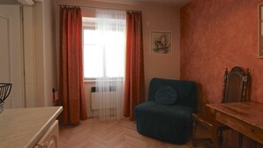 Een kamer met een houten visgraatvloer, een blauwe fauteuil, een houten tafel en een raam met dunne witte gordijnen en oranje gordijnen met een patroon.
