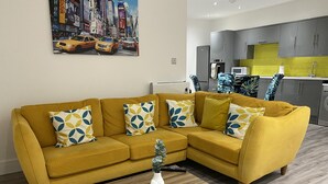 Un soggiorno con un divano color senape, cuscini decorati, un vaso bianco e un quadro che raffigura una strada cittadina.