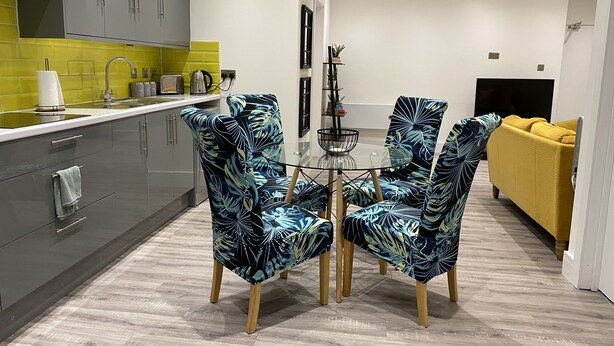 Una cucina moderna con mobili grigi, un pannello di divisa giallo e un'area pranzo con tavolo e sedie decorate con motivi a foglia tropicale.
