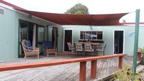 Een overdekt terras buiten met een houten dek, rieten stoelen, een tafel met stoelen en een bordje met de tekst 'Land's End'.