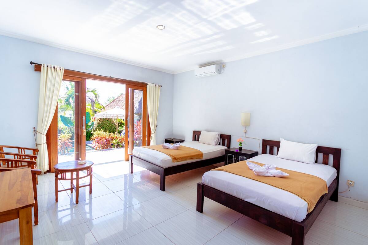 A hotel room with two beds, a wooden desk, and a view of a garden through a glass door.