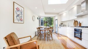 A modern kitchen with white cabinets, a dining area with wooden chairs, and a large window providing a view of the outdoors. - Family home in the heart of Paddington (Paddington)