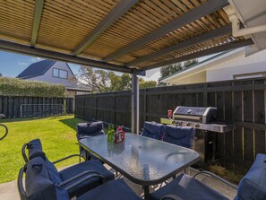 An outdoor patio with a glass table, blue cushioned chairs, a grill, and a covered area with a wooden ceiling. - Walk to the Water - Whangamata Holiday Home (Whangamata)