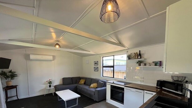 Een compacte woonruimte met een kitchenette, een grijze bank, een klein bijzettafeltje en een hanglamp.