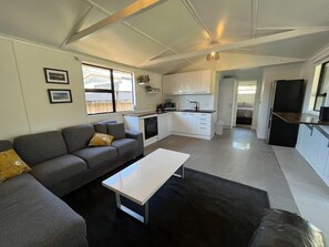 A modern living room with a grey sofa, a white coffee table, and a kitchen area with white cabinets and a black countertop. - Walk to the Water - Whangamata Holiday Home (Whangamata)
