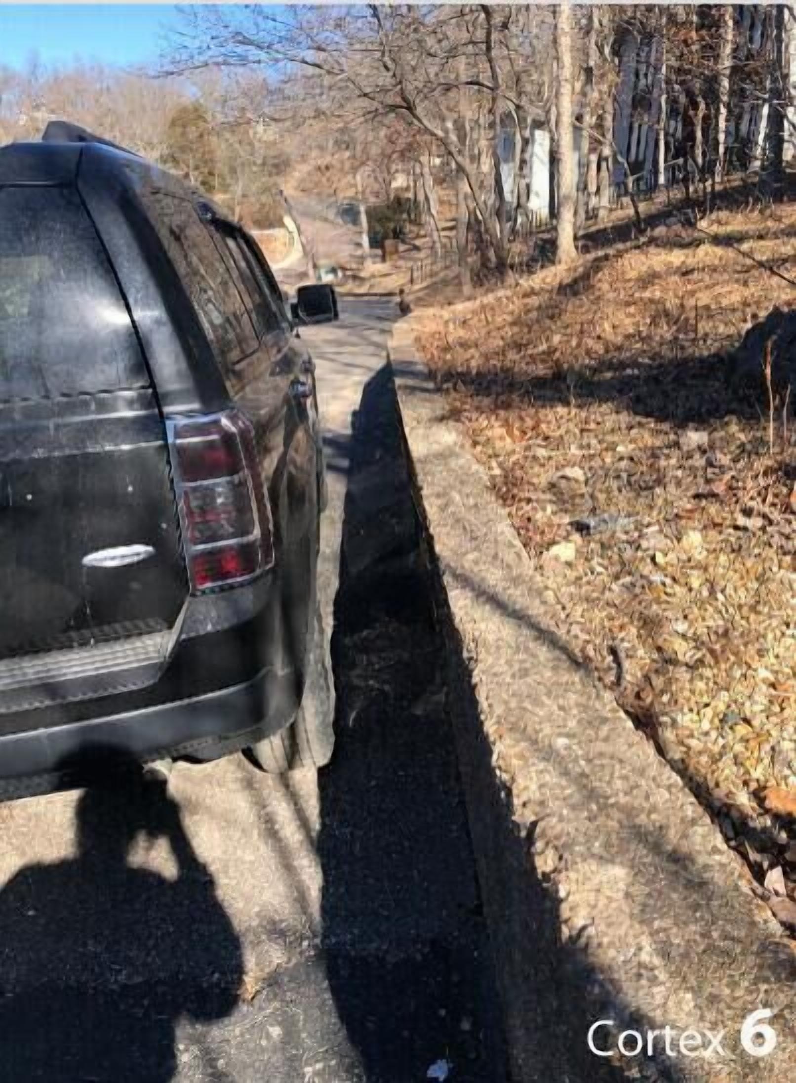 A black SUV parked on a narrow, unpaved road with a curb on one side and a wooded area on the other.