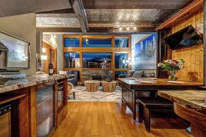A modern kitchen with a stone fireplace, wooden cabinetry, and a large window overlooking a snowy landscape.