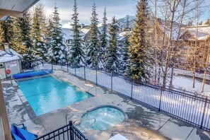 Ein Schwimmbecken mit Whirlpool, umgeben von einem Metallzaun, mit schneebedeckten Bäumen und Bergen im Hintergrund.