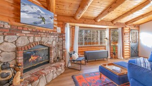 A cozy log cabin interior with a stone fireplace, a large window, and a blue sofa. - Private tree top cabin, Mountain Views, 5 min to slopes, Walk to zoo & food (Big Bear)