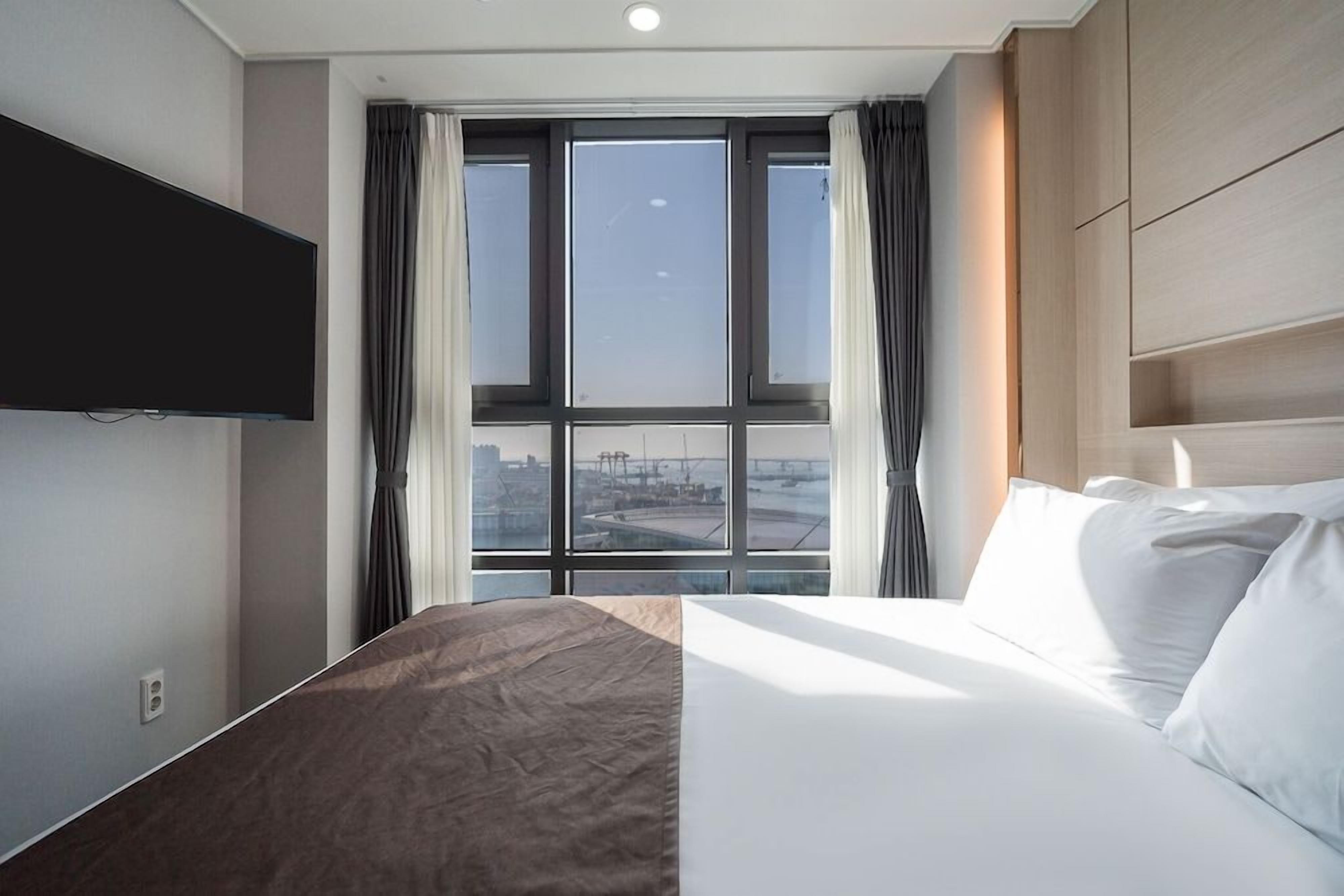 A hotel room with a large bed, a flat-screen TV mounted on the wall, and a window with curtains.