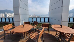 A terrace with wooden tables and chairs, offering a view of a cityscape and a body of water. - Griffin Bay Hotel (Busan)