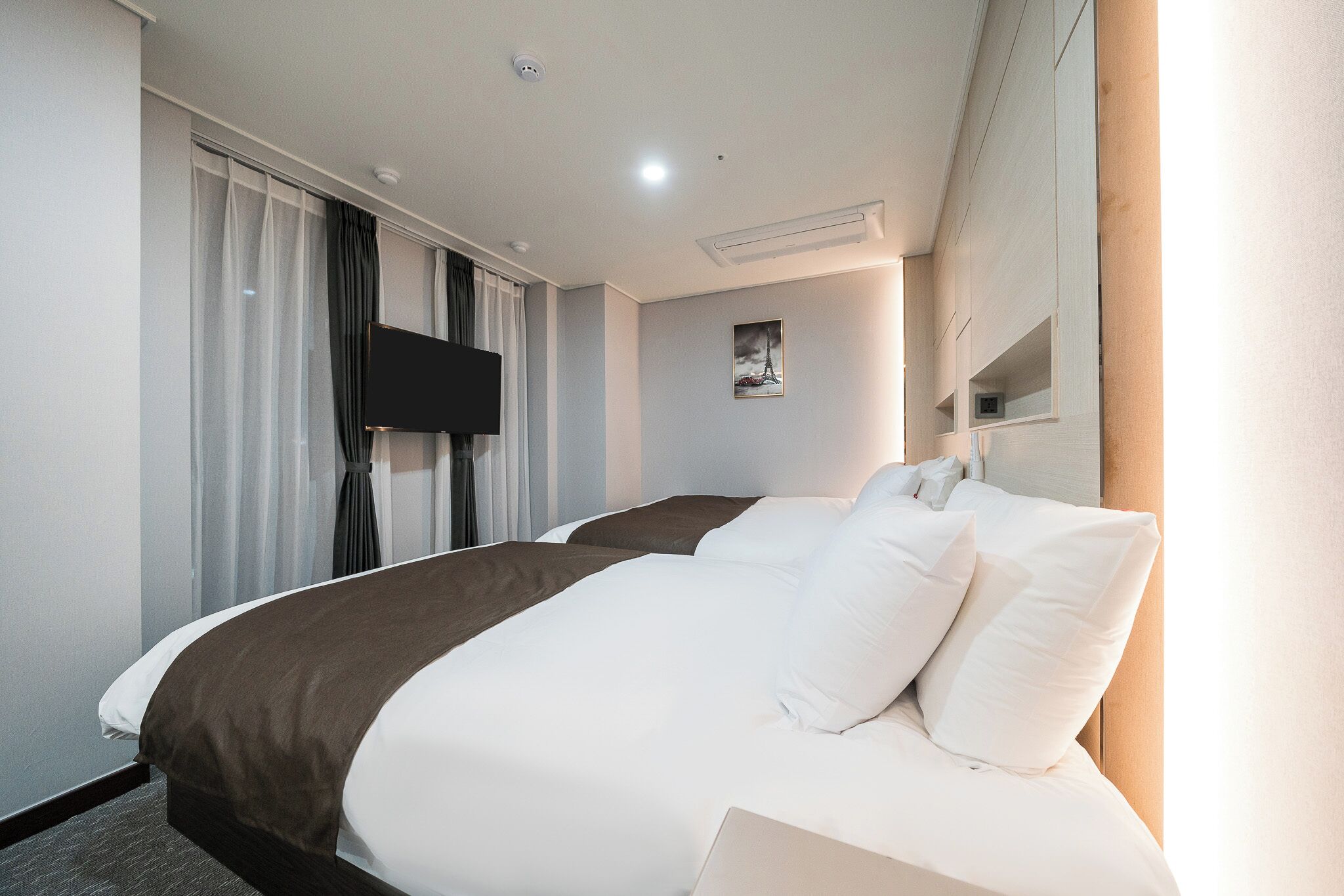 A hotel room with two beds, a flat-screen TV, a painting on the wall, and a window with curtains.