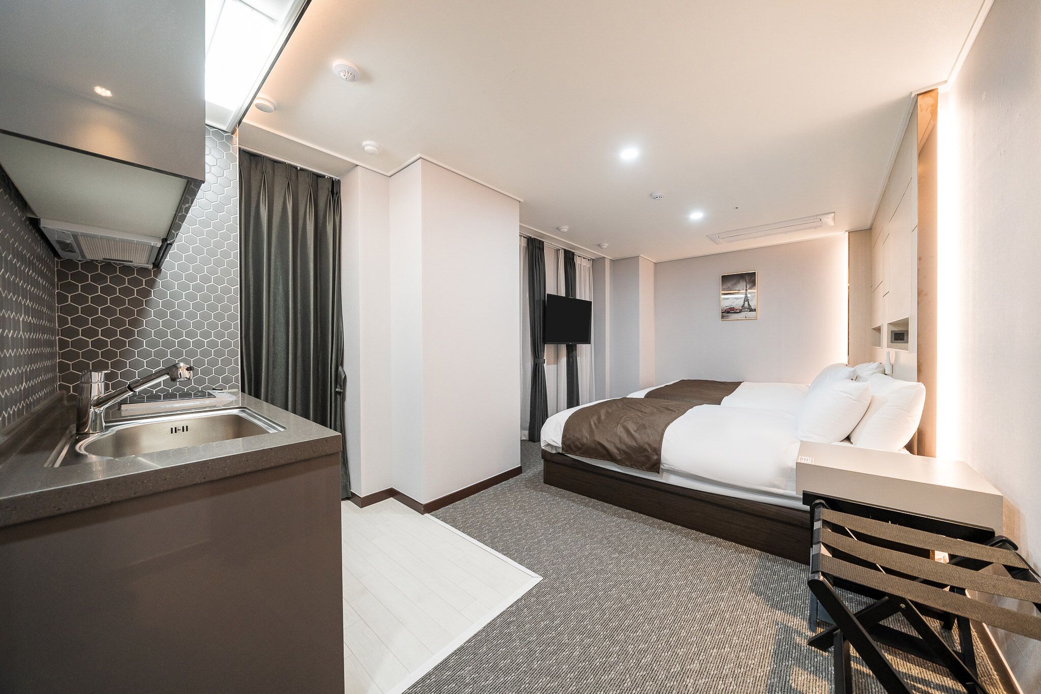 Una habitación de hotel moderna con kitchenette, una cama con una manta marrón, un televisor de pantalla plana y una pequeña imagen de la Torre Eiffel en la pared.