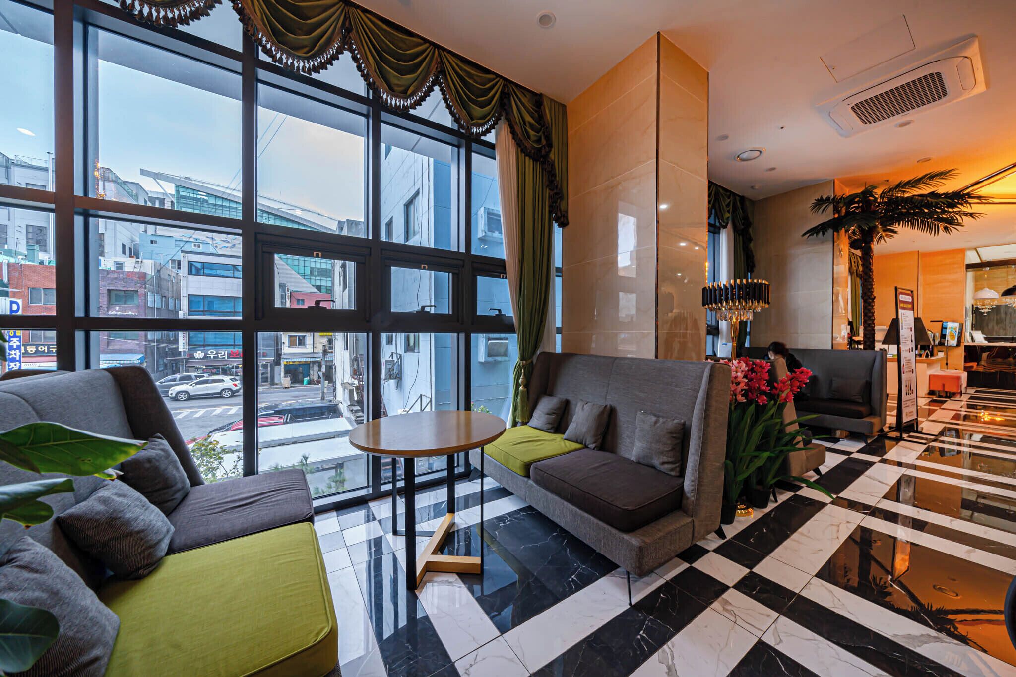 Um saguão de hotel com piso xadrez, um sofá, uma mesinha e uma janela grande com cortinas.