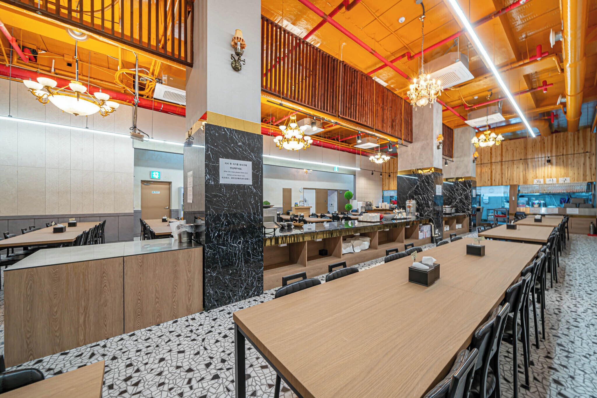 A modern restaurant with long communal tables, high ceilings with exposed beams, and pendant lighting.