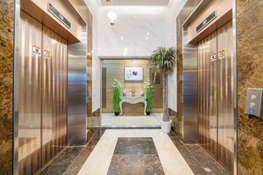 A modern elevator lobby with marble flooring, two elevators, a reception desk, and potted plants. - Griffin Bay Hotel (Busan)