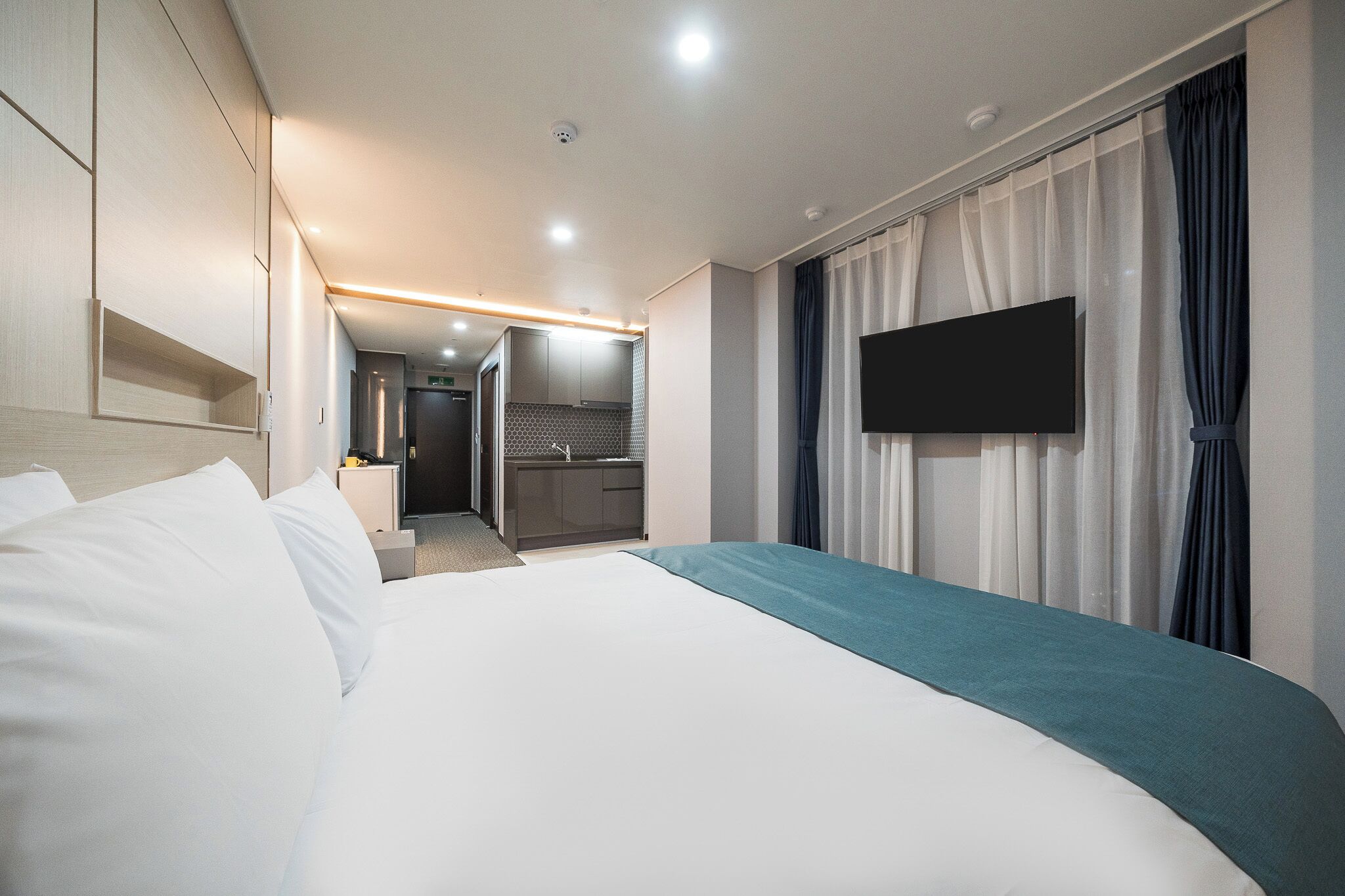 Habitación de hotel con una cama grande, un televisor de pantalla plana en la pared, una kitchenette al fondo y la puerta del baño.