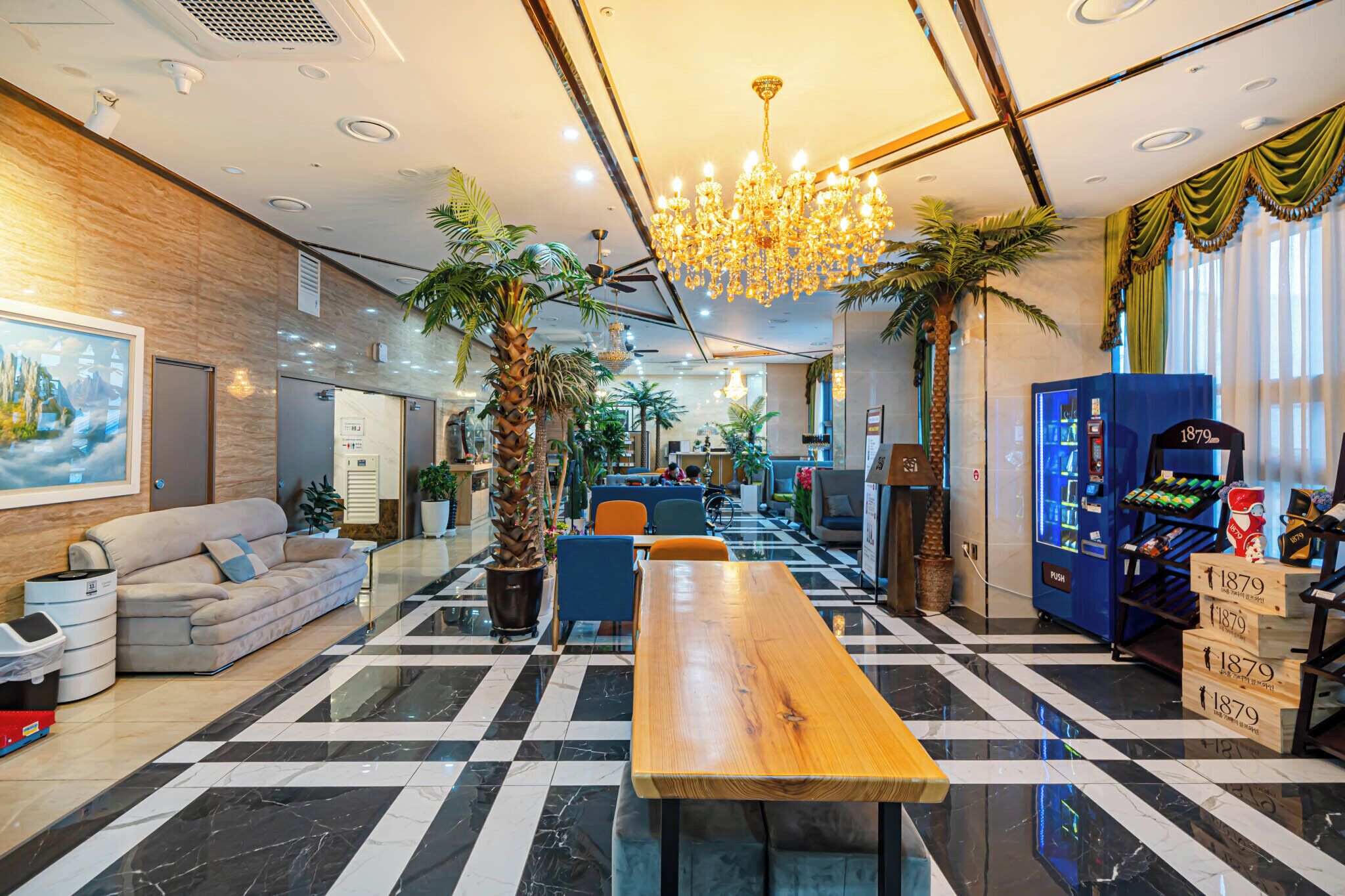 A hotel lobby with a checkered floor, a wooden reception desk, a chandelier, and a vending machine.
