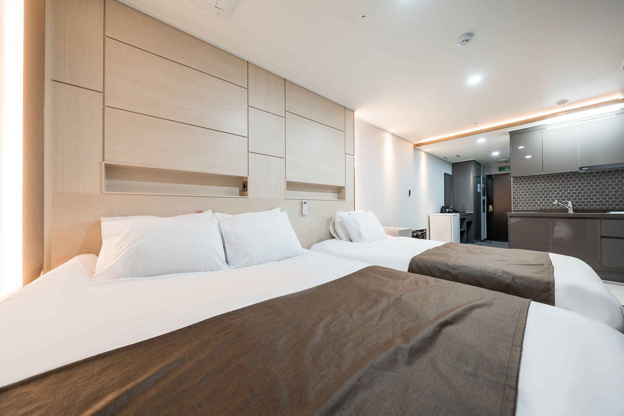A hotel room with two beds, a wooden headboard, a kitchenette, and a bathroom.
