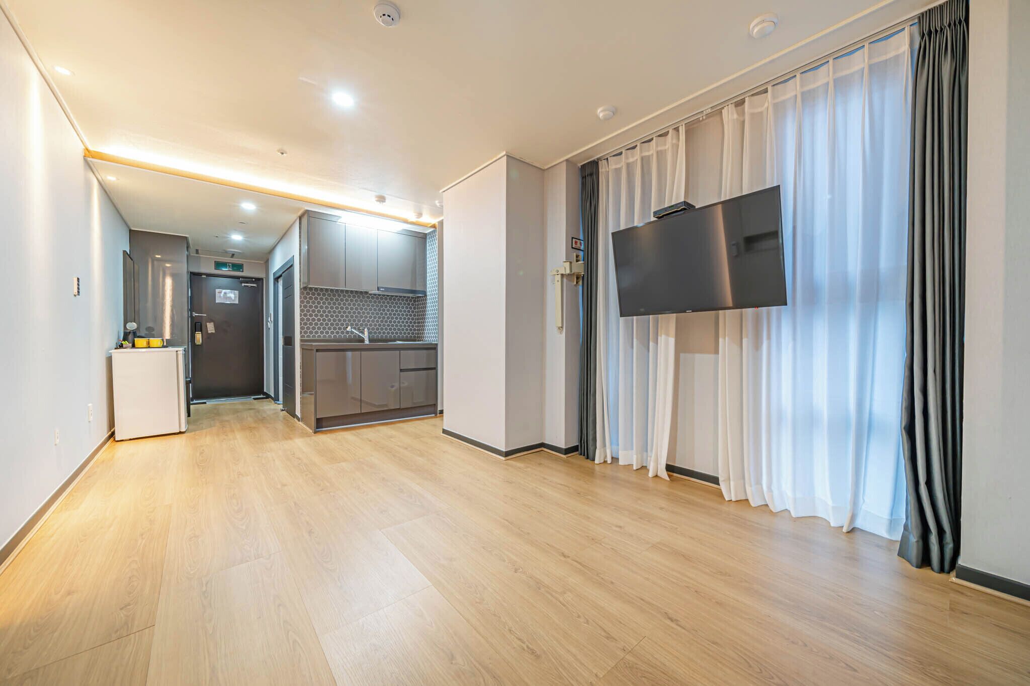 A modern hotel room with a wooden floor, a flat-screen TV mounted on the wall, and a kitchen area visible through an open door.