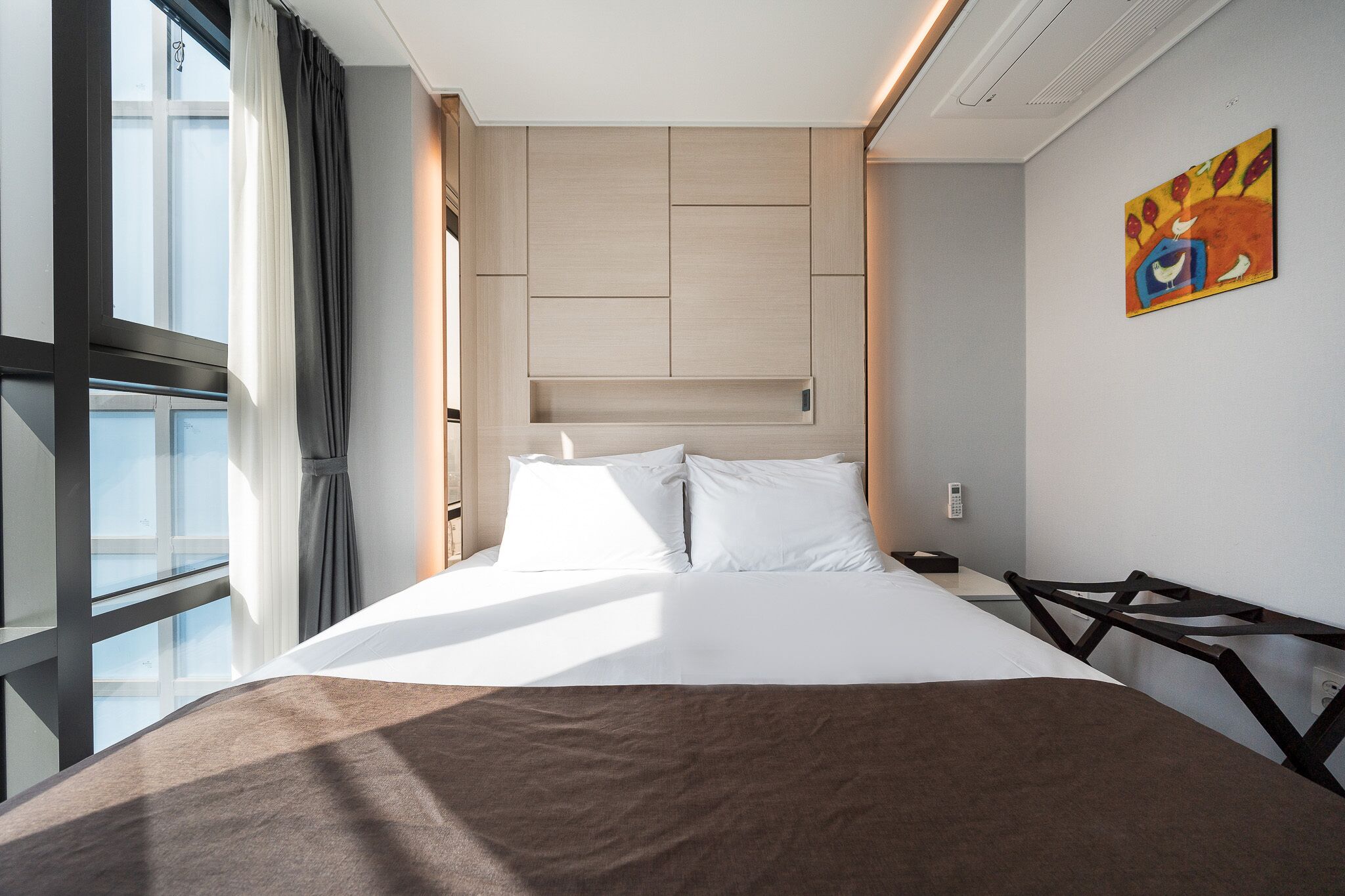 A hotel room with a large bed, a bedside table, a chair, a window with curtains, and a painting on the wall.