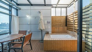 A modern room with a wooden deck, a seating area with a table and chairs, a wooden sauna, and a large window offering a city view. - Griffin Bay Hotel (Busan)