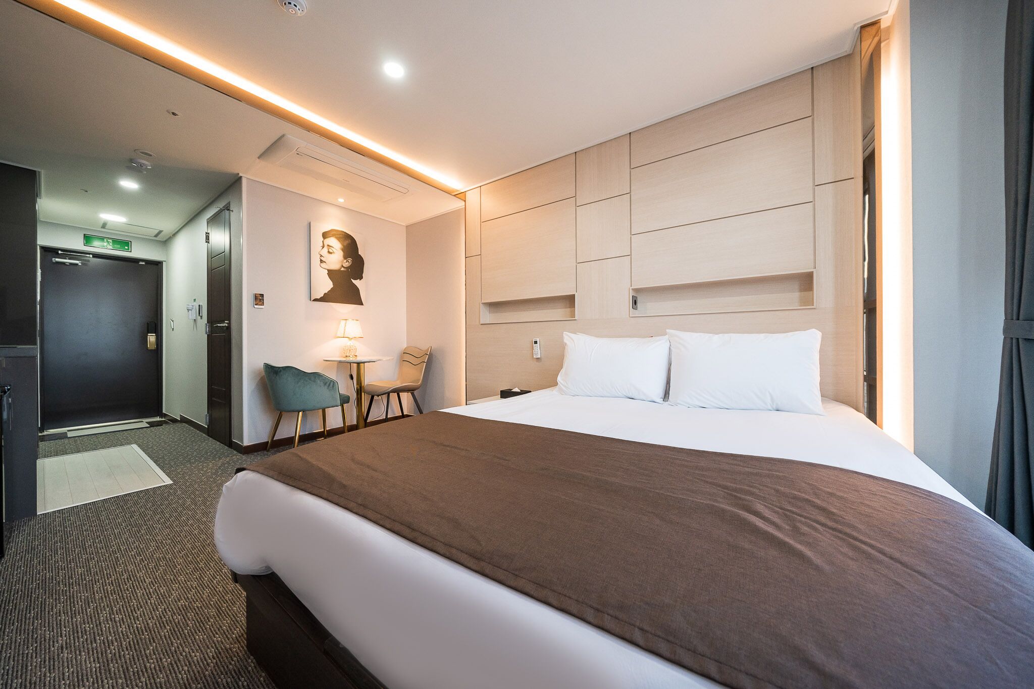 Habitación de hotel con una cama grande, un escritorio con silla, una lámpara de pared y una puerta que da a un pasillo.