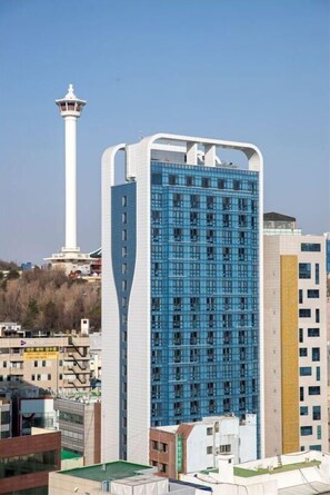 A modern building with a unique curved facade, a prominent tower, and a mix of residential and commercial structures in the background. - Griffin Bay Hotel (Busan)