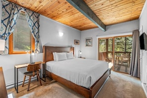 A bedroom with a wooden ceiling, a large bed, a desk with a chair, a window with curtains, and a view of trees through a sliding door.