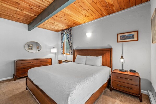 A bedroom with a wooden ceiling, a bed with white bedding, a nightstand with a lamp, and a wall-mounted picture.