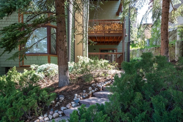 A wooden house with a covered porch, surrounded by dense greenery and a stone pathway.