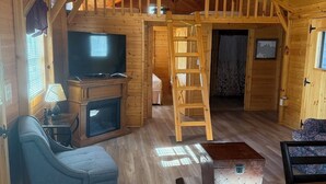 A cozy cabin interior with a wooden ceiling, a television, a fireplace, a ladder, and a dining area.