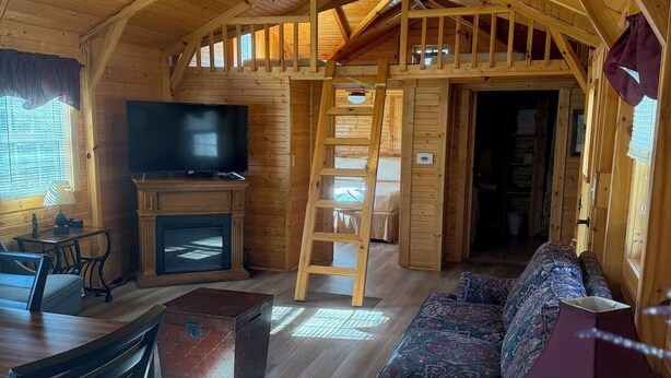 A cozy wooden cabin interior with a TV, sofa, dining table, and a ladder leading to an upper level.