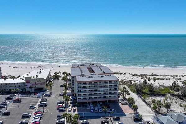 Un hotel frente a la playa con varios pisos, un estacionamiento y una playa al fondo.