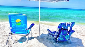 Zwei Liegestühle und ein Sonnenschirm an einem Sandstrand mit klarem, türkisblauem Wasser.