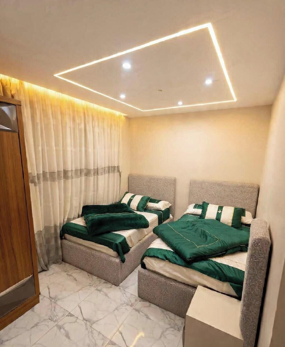 A hotel room with two beds, a wooden wardrobe, a marble floor, and a modern ceiling light fixture.