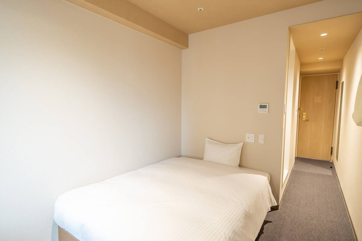 A small, single-bed room with a plain white wall, a wooden door, and a carpeted floor.