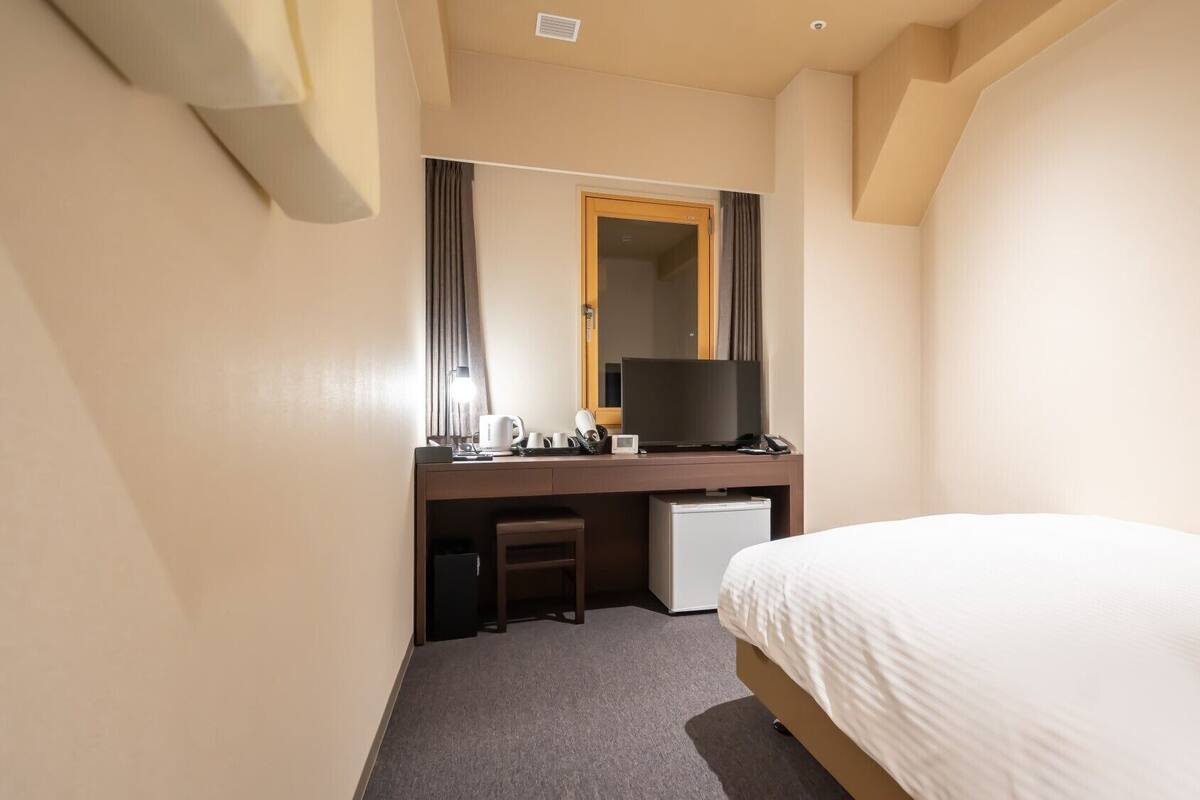A hotel room with a bed, a desk with a television, a small refrigerator, and a mirror.